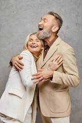 A mature, elegant couple dressed in debonair attires enveloped in a warm, loving embrace against a gray backdrop.