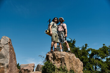 Two young men, a gay couple, hike together in nature on a summer day. They stand on a rocky...