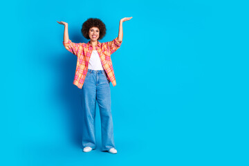 Full size photo of attractive young woman raise hands hold empty space dressed stylish checkered clothes isolated on blue color background