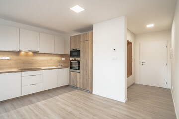 A modern kitchen with white cabinets, wooden countertops, built-in appliances, under-cabinet lighting, light wooden flooring, an entryway with a white door, and a wooden bench.