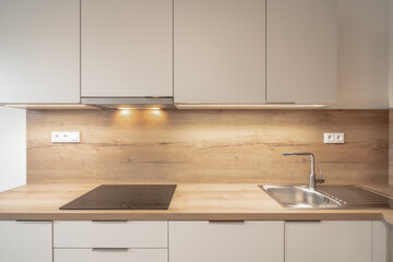 A modern kitchen with a wooden backsplash, white cabinets, under-cabinet lighting, an electric cooktop, and a stainless steel sink.