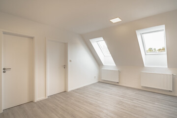 
A bright, minimalist room with a slanted ceiling, two skylight windows, white walls, two radiators, and two closed white doors.