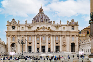 Rome, Italy. The front facade, dome, and colonnade of Saint Peter's Basilica dominate the scene, while the bustling square below is filled with crowds of people and tourists.