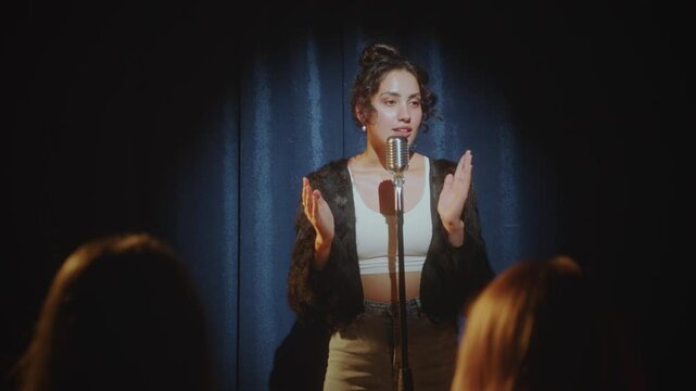 Young female stand-up comedian standing on stage in a spotlight and performing monologue in mic. View from audience