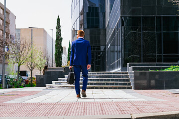 A man in a blue suit walks down a sidewalk with a briefcase in his hand