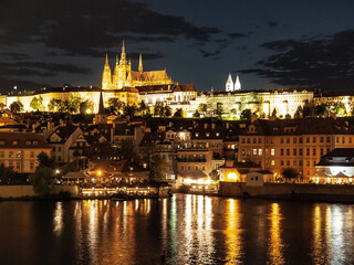 Obraz premium Prague Castle illuminated at night, with the Vltava River reflecting the city lights. Buildings and spires are visible against the dark sky.