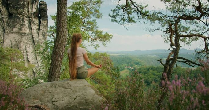 Woman in a meditative pose on a cliffside overlooking a vast green forest in summer day