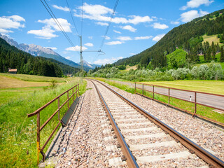 In the Dolomites, Italy, a set of train tracks runs through a scenic valley surrounded by green fields, forests, and snow-capped mountains under a bright blue sky.