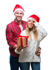 Young couple in love wearing christmas hat and holding present over isolated background with happy face smiling doing ok sign with hand on eye looking through fingers