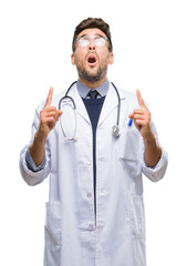 Young handsome doctor man over isolated background amazed and surprised looking up and pointing with fingers and raised arms.