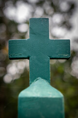 Close-up photo of a Christian cross on a cemetery iron green fence against a blurred background in a natural ambiance with bokeh effect