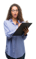 Young hispanic business woman holding clipboard scared in shock with a surprise face, afraid and excited with fear expression