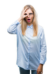 Young beautiful blonde business woman wearing glasses over isolated background doing ok gesture shocked with surprised face, eye looking through fingers. Unbelieving expression.