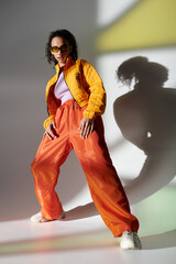 A stylish young woman poses confidently in a yellow jacket and orange pants, her sunglasses adding an edge to her look.