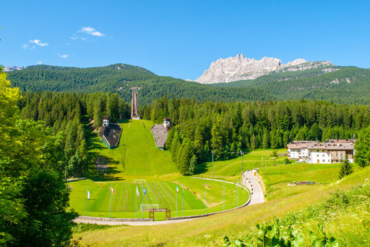 A view of the ski jump hill, Trampolino Olimpico Italia, in Cortina d'Ampezzo, Italy, surrounded by lush green trees and mountains.