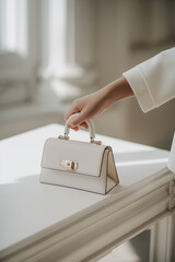 Naklejka premium A close-up shot of a woman's hand reaching for an elegant handbag on a minimalist white table under soft natural light.