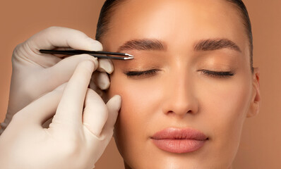 Hand holding tweezers and shaping eyebrows to young European woman, beautician plucking brow hairs...
