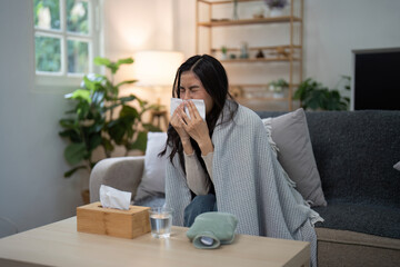 Woman Sneezing into Tissue While Sitting on Sofa at Home, Surrounded by Plants and Blankets,...