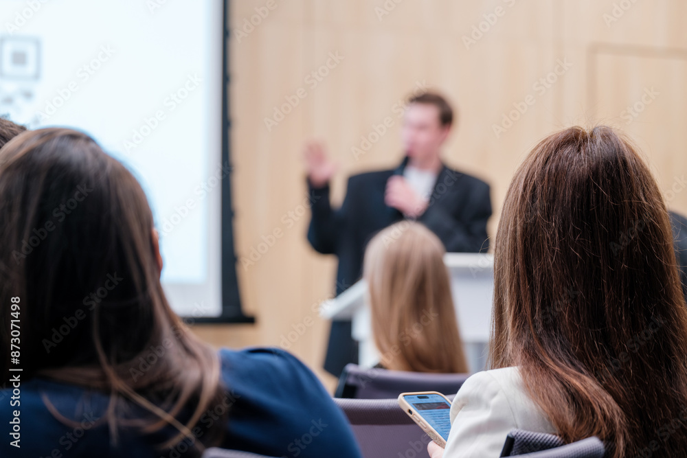 Wall mural People in an audience listening to a business presentation with a blurry speaker in the background. Professional setting during a conference or seminar.