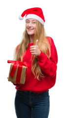 Blonde teenager woman wearing santa claus hat happy with big smile doing ok sign, thumb up with fingers, excellent sign