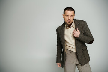 A stylish man in a tweed blazer and khaki pants poses against a grey background.