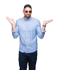 Young handsome man wearing sunglasses over isolated background clueless and confused expression with arms and hands raised. Doubt concept.