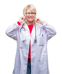 Young beautiful blonde doctor woman wearing medical uniform over isolated background covering ears with fingers with annoyed expression for the noise of loud music. Deaf concept.