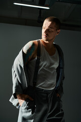 Man with buzz cut and tattoos in studio, wearing white tank top, suspenders, gray jacket.