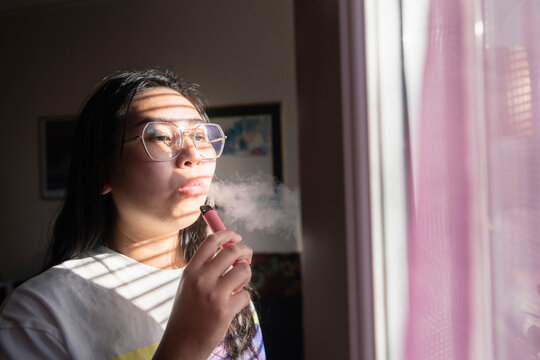 Young woman smoking vape pen while standing near window