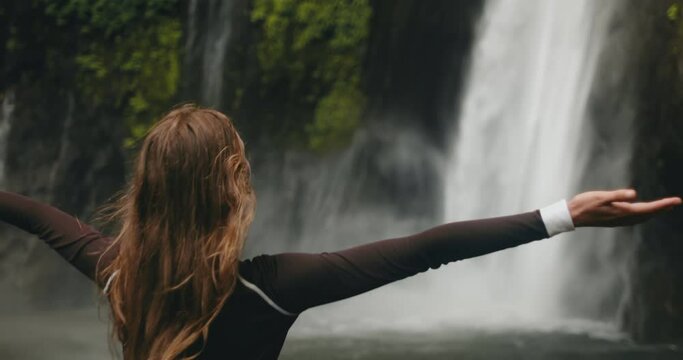 Traveler hiking girl raising arms standing at powerful waterfall Air Terjun Munduk, Indonesia, Bali island. Woman posing, back view, slow motion. Beautiful nature landscape. Travel, tourism, holiday
