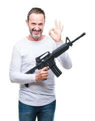 Middle age senior hoary criminal man holding gun weapon over isolated background doing ok sign with fingers, excellent symbol