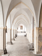 A long, arched walkway with white stone columns leads to a courtyard with a bench in the distance.