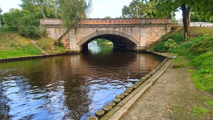 The high grassy banks of the river flowing in the city park are reinforced with logs. There are trees growing all around. A stone bridge stands over the river. Clear summer weather