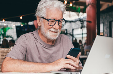Attractive senior man sitting in a cafe smiling while using mobile phone. Cheerful senior male, using modern technology expressing happiness and contentment in a relaxed atmosphere