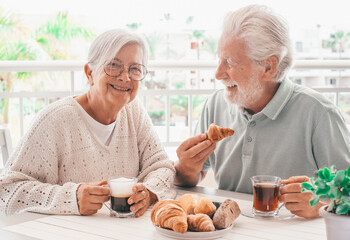 Smiling elderly couple enjoying a morning breakfast together at a cozy outdoor balcony with croissant and coffee and tea, authentic serene retirement lifestyle
