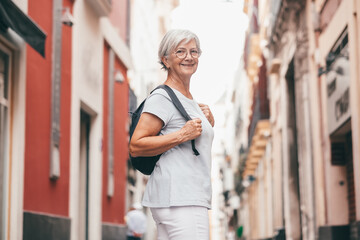Smiling carefree senior woman tourist walking in old town of Seville, Spain holding a backpack enjoying vacation trip freedom, healthy lifestyle in retirement
