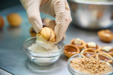 Woman baker dipping sweet pastry nut in white chocolate into coconut flakes
