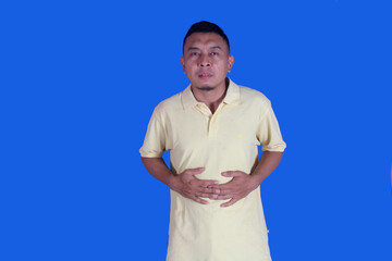 Funny face of Asian man wearing yellow t-shirt suffering from stomach ache due to diarrhea on blue...