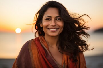 Fototapeta premium Portrait of a grinning indian woman in her 30s wearing a versatile buff over vibrant beach sunset background