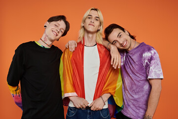 Three young friends in stylish attire pose together with a pride flag draped around one of them,...