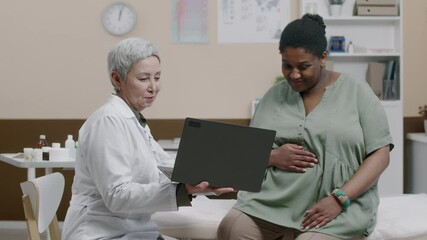 Medium shot of mature Caucasian female gynecologist holding laptop, discussing and reviewing birth plan with young pregnant African American woman caressing belly during appointment