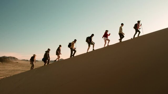 Large group of diverse tourists with backpacks and hiking poles goes uphill on sand dune against sunset sky in desert