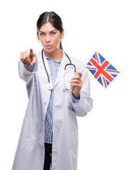 Young hispanic doctor woman holding flag of united kingdom pointing with finger to the camera and to you, hand sign, positive and confident gesture from the front