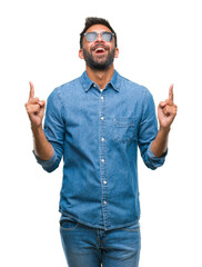 Fototapeta premium Adult hispanic man wearing sunglasses over isolated background amazed and surprised looking up and pointing with fingers and raised arms.