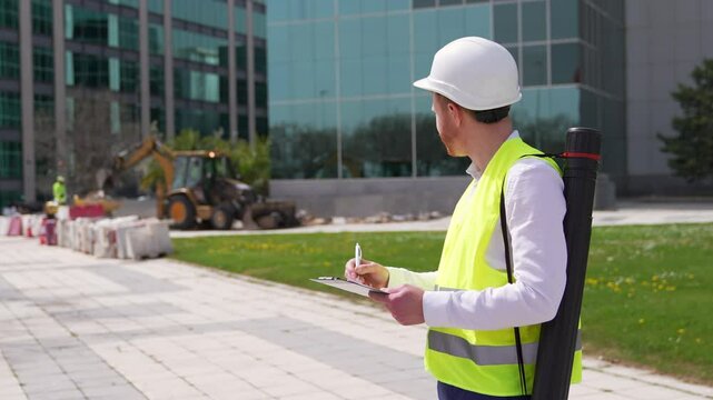 A redheaded quality inspector in a reflective vest and hard hat takes notes while observing construction workers on site.