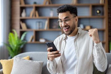 Close-up photo of a happy young Nigerian man at home sitting on the sofa and happy about the news and win, looking at the phone screen and making a yes gesture with his hand