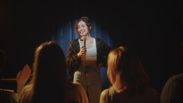 Young female comedian smiling and talking in microphone in front of audience during stand-up performance on stage. Zoom-out shot