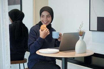 A woman wearing blue sweater is sitting at a table with a laptop and a cup of coffee. She is smiling and she is enjoying her coffee and laptop