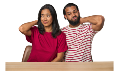 Seated young Latino couple at table touching back of head, thinking and making a choice.