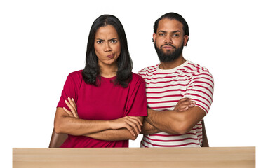 Seated young Latino couple at table frowning face in displeasure, keeps arms folded.